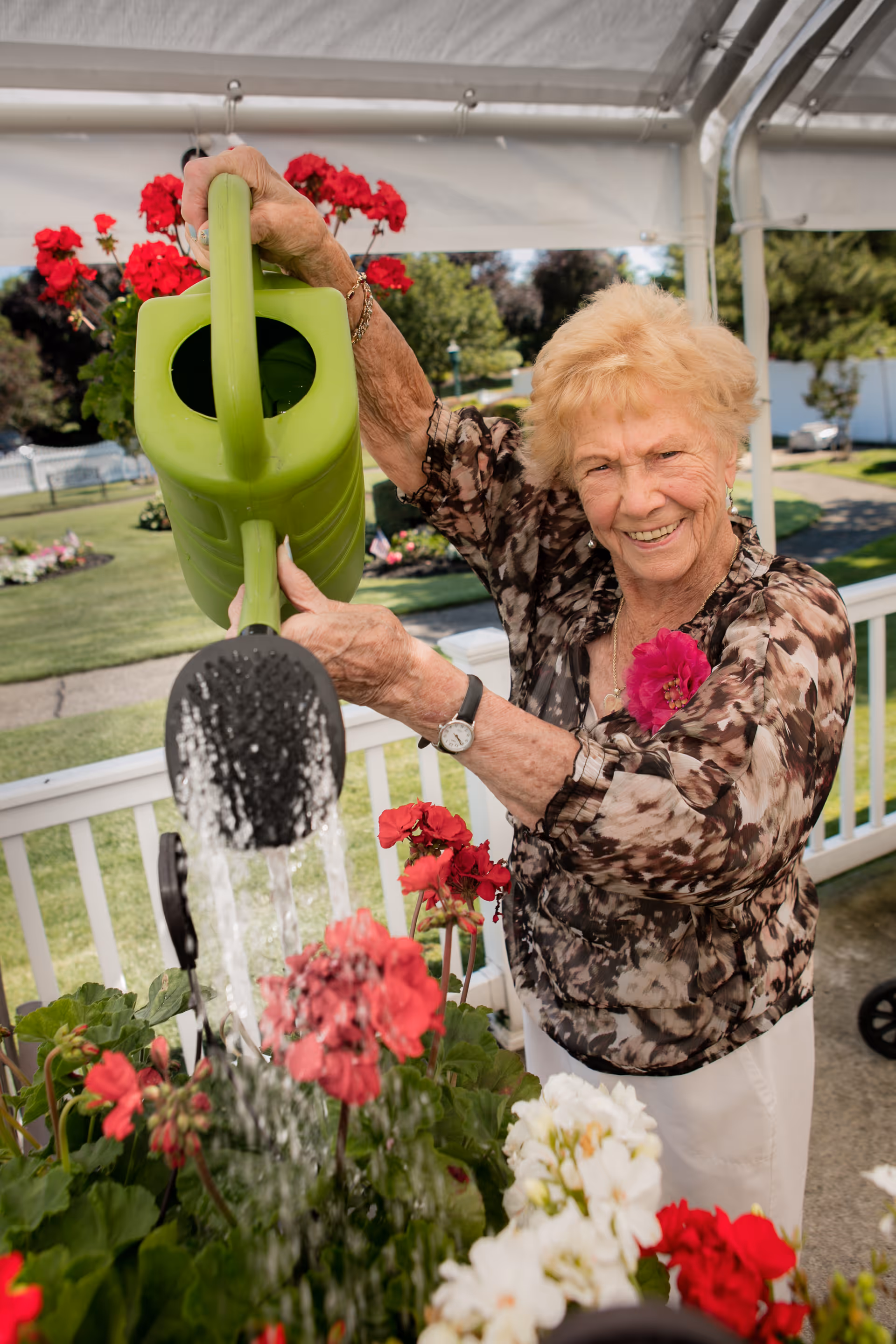 An elderly woman with short blonde hair and a floral blouse is smiling while watering red and white flowers with a green watering can on a porch or patio. There are plants and a garden visible in the background under a white canopy.