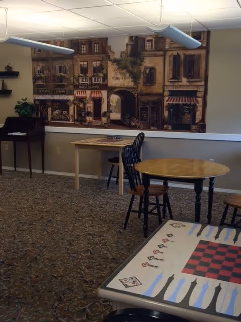 Communal activity room with round and game tables, chairs, a piano, and a large wall mural depicting storefronts.