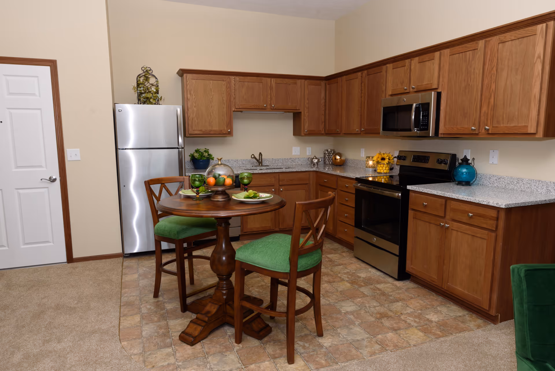 A kitchen area in a retirement community featuring wooden cabinets, a stainless steel refrigerator, stove, and microwave. There is a small round wooden table with two chairs that have green cushions. The floor is a mix of carpet and tile, and there are decorative items on the countertops and table.