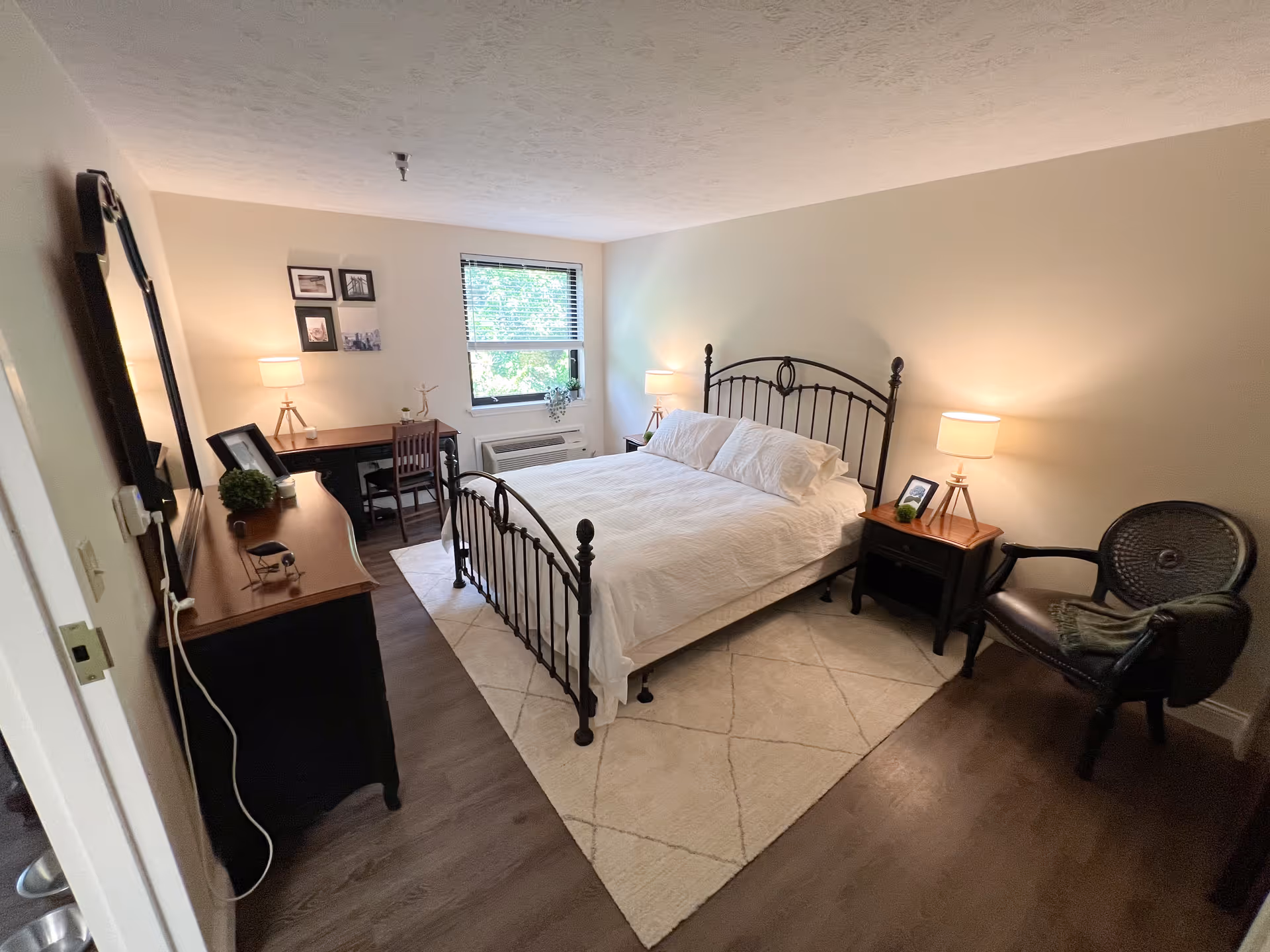 A cozy bedroom in a senior living community featuring a metal-framed bed with white bedding, two bedside tables with lamps, a wooden dresser with a mirror, a desk with a chair near a window, and a comfortable armchair with a blanket. The room has wooden flooring and neutral-colored walls.
