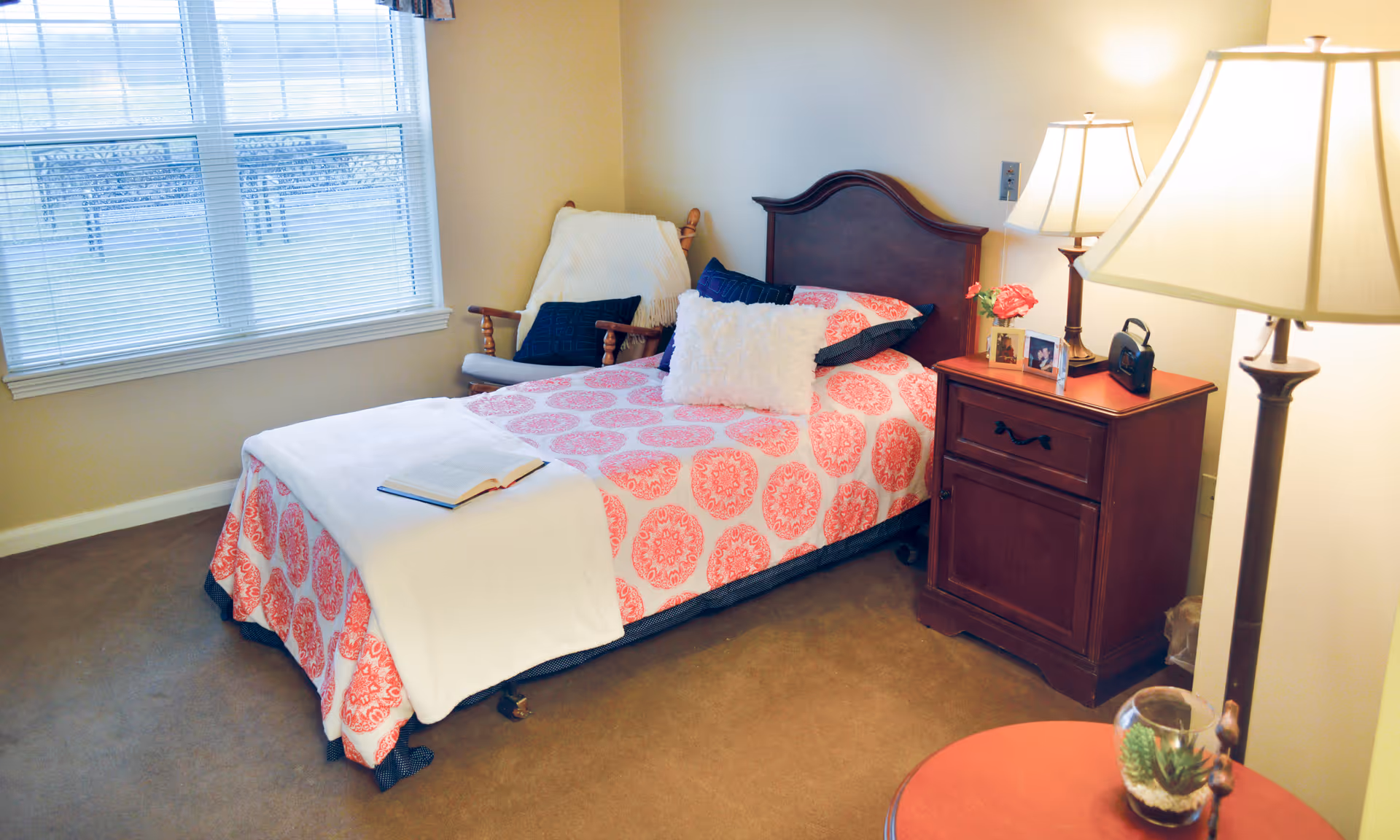 A cozy bedroom with a single bed covered in a pink and white patterned bedspread, a white blanket, and several pillows. Next to the bed is a wooden nightstand with a lamp, framed photos, and a small flower vase. A wooden rocking chair with cushions is placed near a window with blinds. The room has beige walls and carpeted floor.