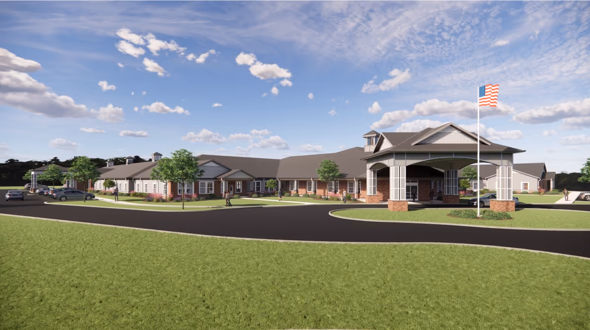 Wide exterior view of a single-story assisted living facility with a covered porte-cochère, circular driveway, flagpole, and landscaped lawns under a blue sky.
