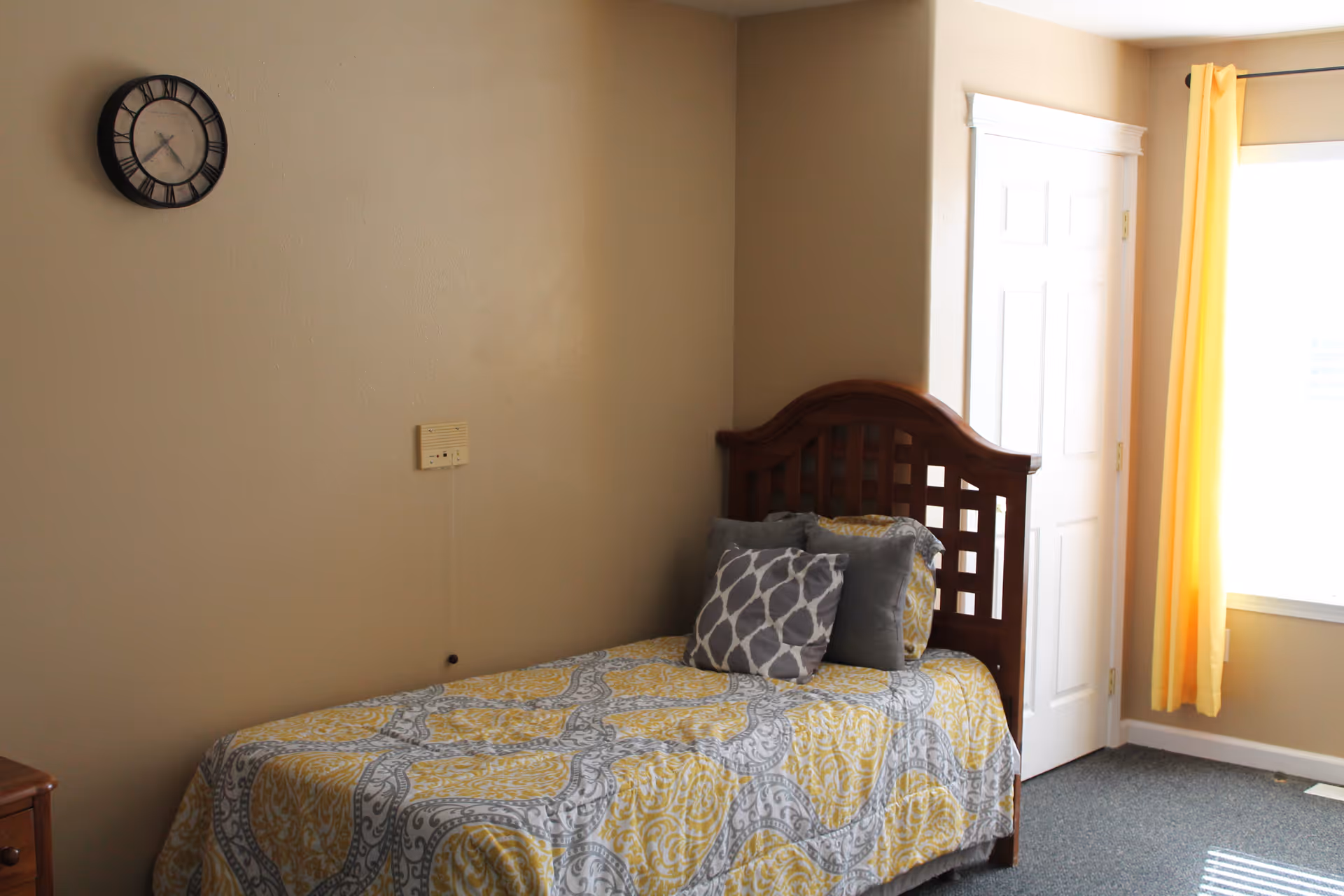 A small bedroom with a single wooden bed featuring a yellow and gray patterned bedspread and several pillows. There is a wall clock above the bed, a closed white door, and a window with yellow curtains letting in natural light.