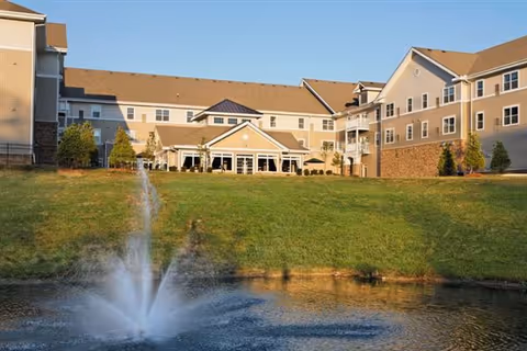 Exterior view of a large senior living facility named Pinebrook with beige siding and a brown roof, situated behind a grassy hill and a pond with a water fountain spraying water upwards.