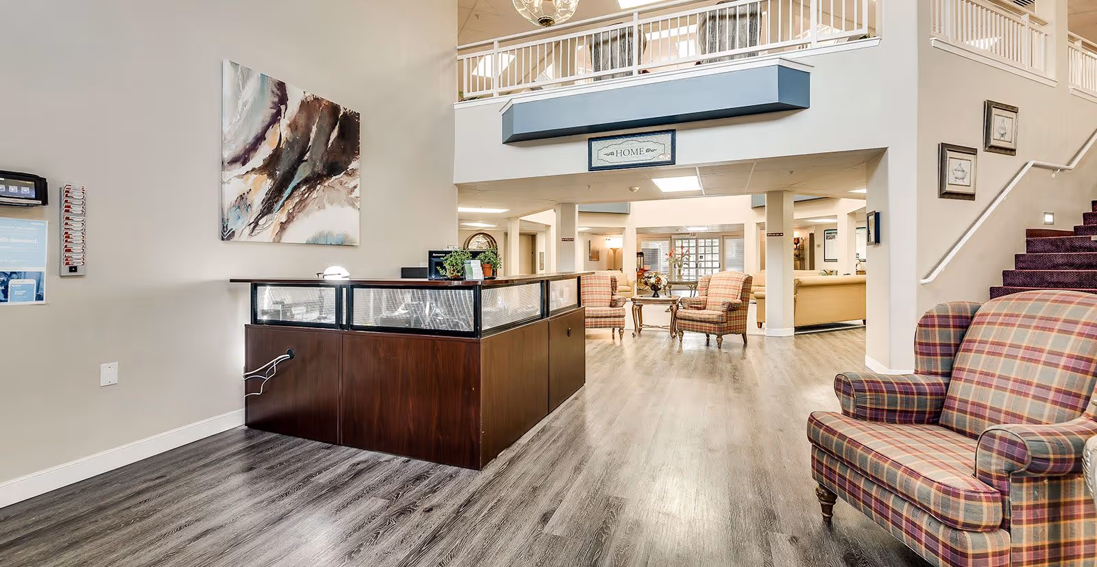 Interior view of a senior living facility lobby with a reception desk on the left, plaid upholstered armchairs on the right and in the background, light wood flooring, neutral-colored walls, and a staircase with a purple carpet leading upstairs. There is a large abstract painting on the wall and a sign above the reception area that reads 'HOME'.