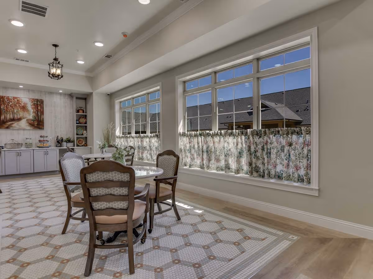 A bright dining area with large windows covered halfway by floral curtains, a round table with four cushioned wooden chairs, a tiled floor with a geometric pattern, and a sideboard with decorative plates and a painting of a tree-lined path on the wall.