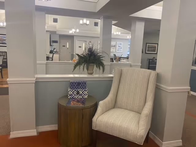 A cozy seating area in a senior living facility featuring a beige upholstered armchair next to a round wooden side table with a decorative plant and a patterned book on top. The background shows a spacious, well-lit interior with columns, mirrors, and additional seating areas.