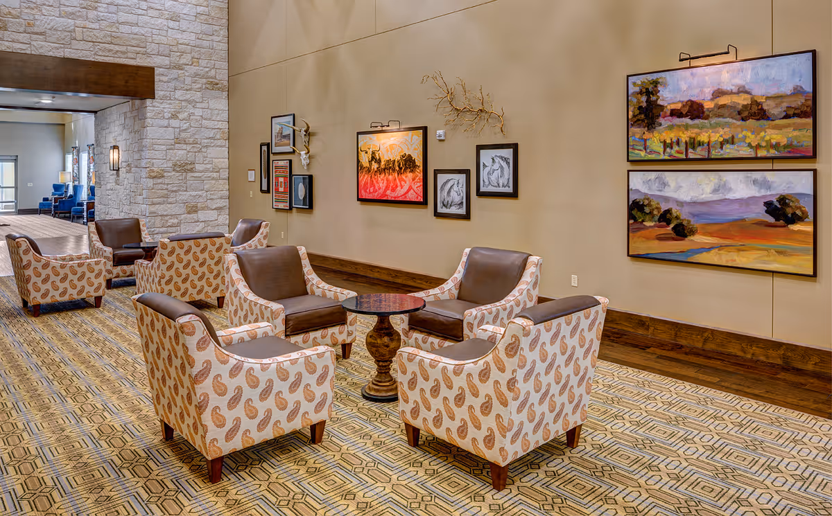 A cozy seating area in a senior living facility with four patterned armchairs arranged around a small round wooden table. The walls are decorated with various framed paintings and artwork, and the floor has a geometric patterned carpet. The space has a warm and inviting atmosphere with a stone wall and additional seating visible in the background.