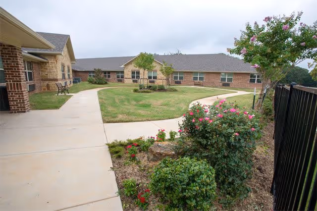 Outdoor courtyard area at Canton Oaks featuring a curved concrete walkway, green lawn, flowering bushes, and single-story brick buildings with windows in the background under a cloudy sky.