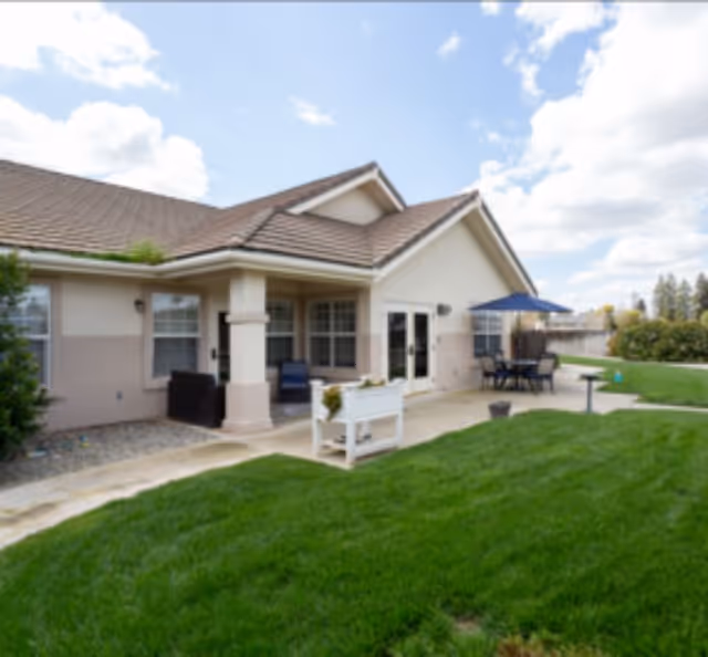 Outdoor patio area of a single-story building with beige walls and a brown tiled roof. The patio has seating including chairs and a table with a blue umbrella. There is a well-maintained green lawn in the foreground and a partly cloudy sky above.
