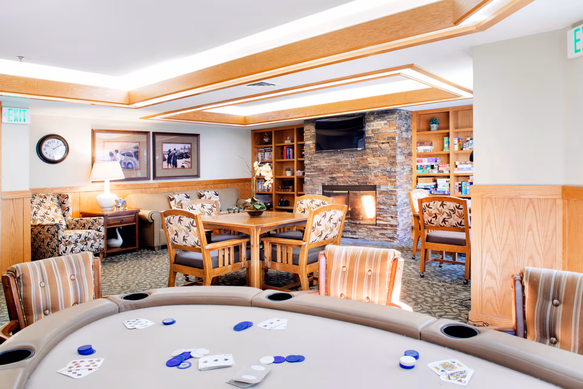 Bright communal lounge with a poker table in the foreground, seating, bookshelves and a stone fireplace with TV in the background.