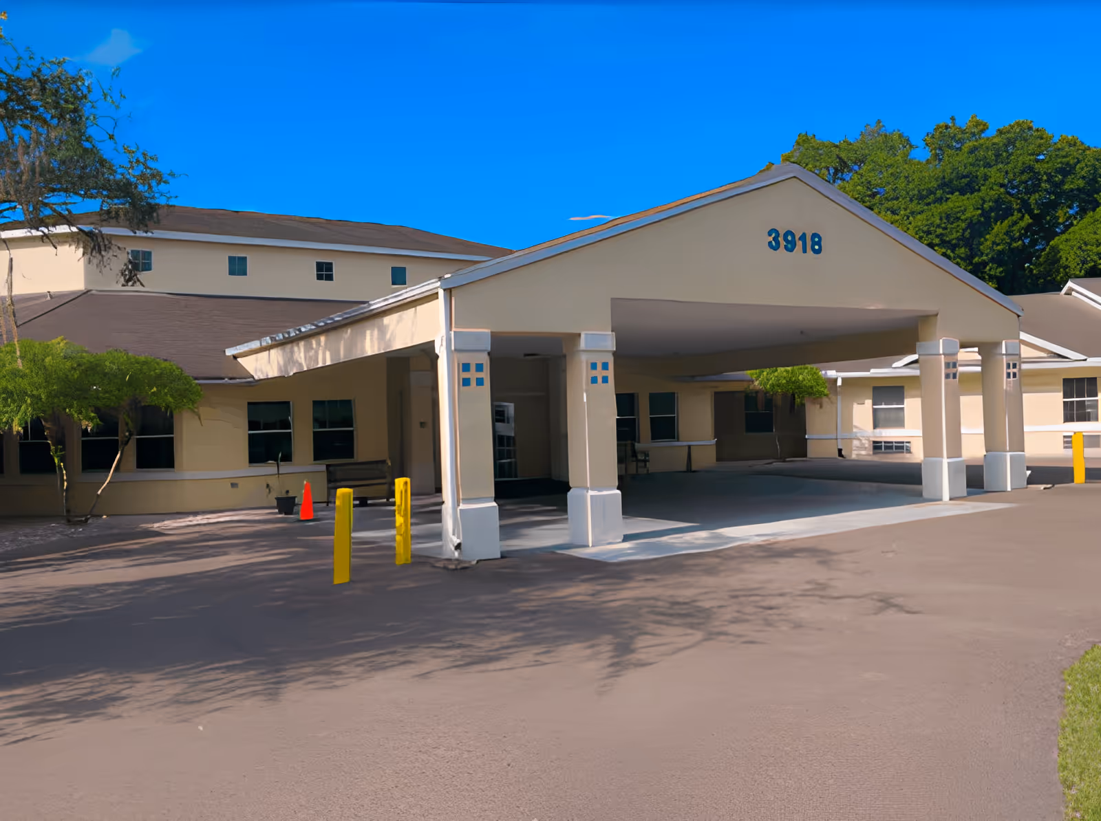 Exterior view of a single-story building with a covered entrance supported by columns. The building is beige with a brown roof and has the number 3918 displayed on the front of the covered entrance. There are trees and bushes around the building and a clear blue sky above.