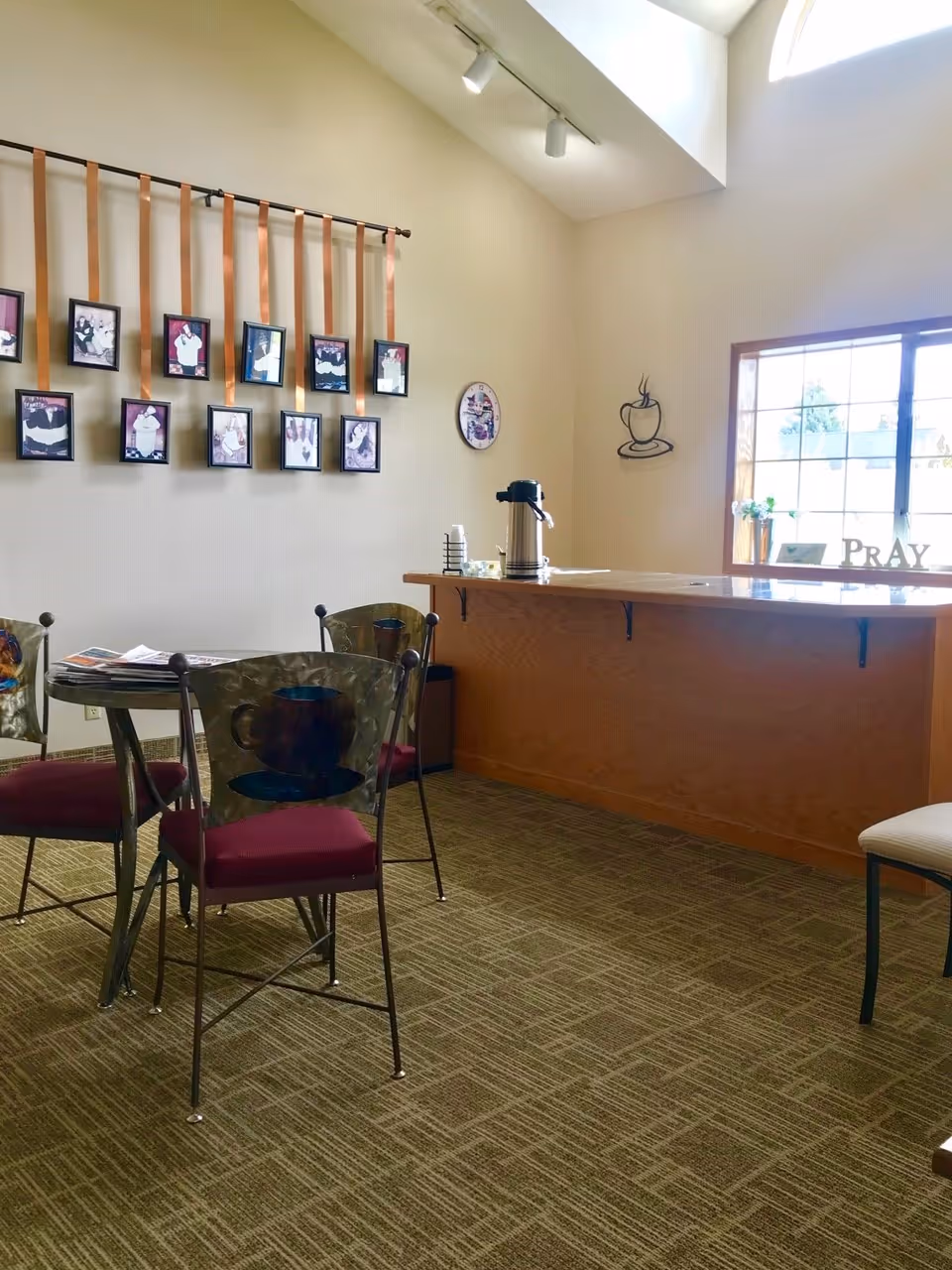 A cozy seating area with a round table and four metal chairs with maroon cushions and coffee cup designs on the backrests. On the left wall, there are several framed pictures hanging from orange ribbons. A wooden counter with a coffee dispenser and cups is positioned near a window letting in natural light. The wall has coffee-themed decorations and a sign that says 'PRAY'.