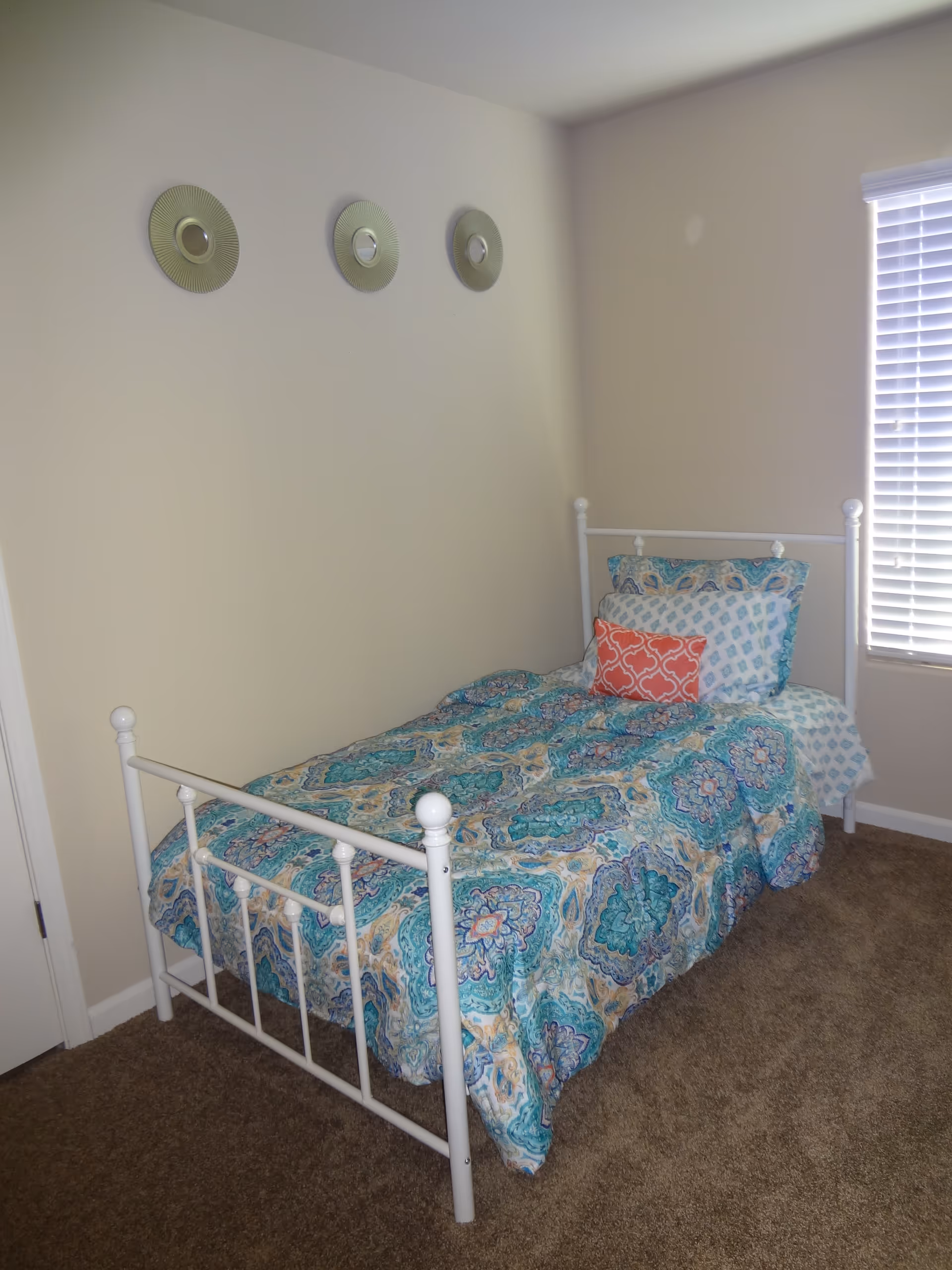A small bedroom with a single white metal bed frame. The bed is made with a colorful blue, teal, and beige patterned comforter and multiple pillows, including one coral-colored decorative pillow. The room has beige walls with three round decorative mirrors hanging above the bed. There is a window with white blinds on the right side and brown carpeted flooring.