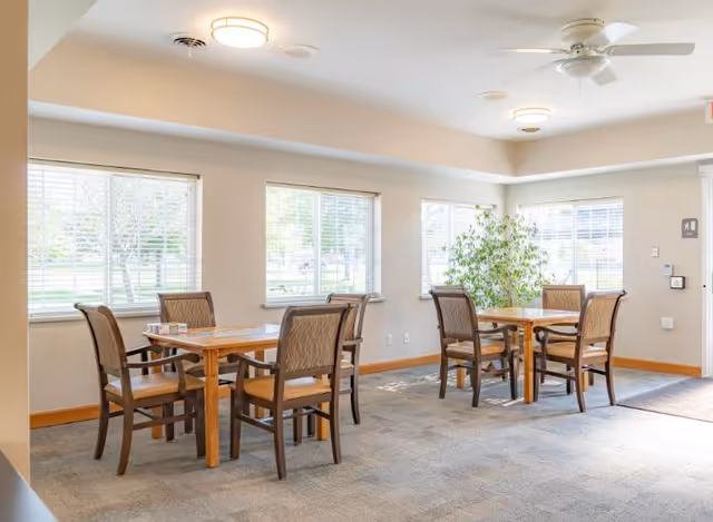Bright communal dining area with two wooden tables and chairs, large windows, and a potted plant.