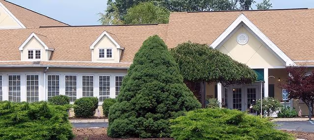 Exterior view of a single-story building with a tan roof and multiple white-framed windows. The entrance has a small peaked roof with a green awning. The foreground features well-maintained landscaping with various green shrubs and trees.