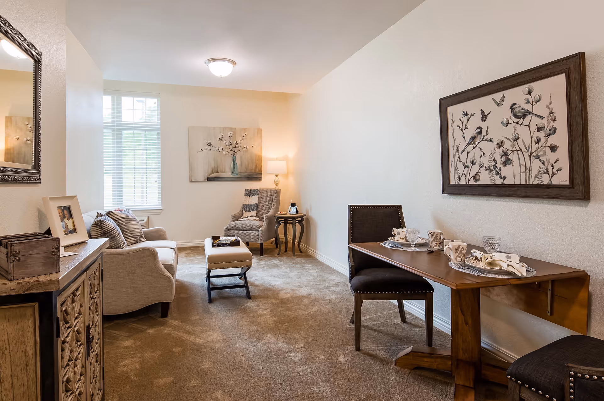 A cozy living area with a beige sofa, a cushioned armchair, and a small ottoman. There is a wooden side table with a lamp and a framed picture on a wooden cabinet. A wooden dining table with two chairs is set with plates, cups, and napkins. The walls are decorated with framed artwork, and a window lets in natural light.
