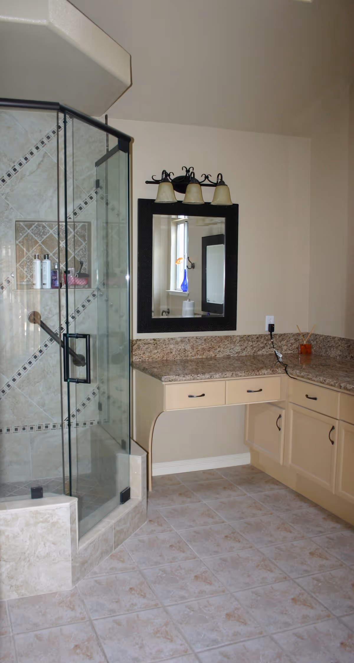 A bathroom with a glass-enclosed shower featuring tiled walls with decorative accents. There is a granite countertop with beige cabinets underneath, a black-framed mirror above the counter, and a three-light fixture mounted above the mirror. The floor is tiled with light-colored tiles.