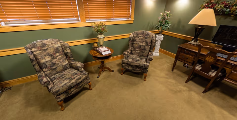 A cozy room with two patterned armchairs placed around a small wooden round table holding a decorative globe and a book. The room has green walls with wooden trim, a window with closed blinds, a wooden desk with a lamp and computer, and a plant on a white pedestal in the corner.