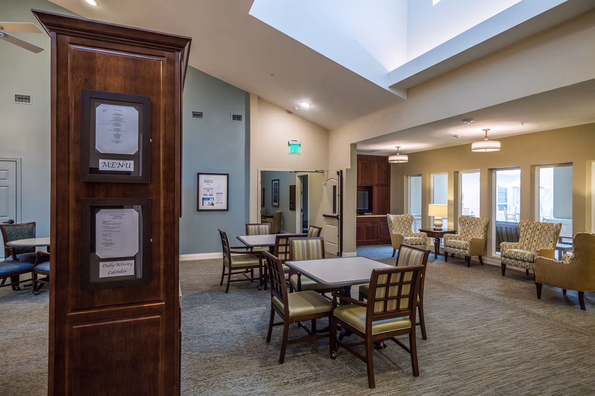 Interior view of a senior living facility common area with tables and chairs for dining or activities, comfortable armchairs arranged near windows, soft lighting from ceiling fixtures and lamps, and a wooden pillar displaying a menu and daily activity calendar.