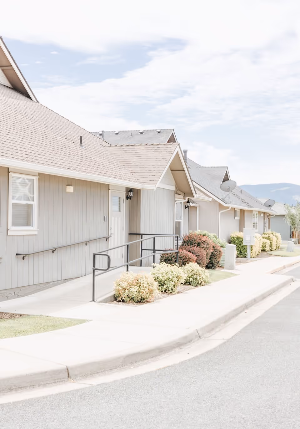Exterior view of a single-story residential building with beige siding, a sloped roof, and a ramp with handrails leading to the entrance. There are small bushes and landscaping along the sidewalk, and mountains are visible in the background under a partly cloudy sky.