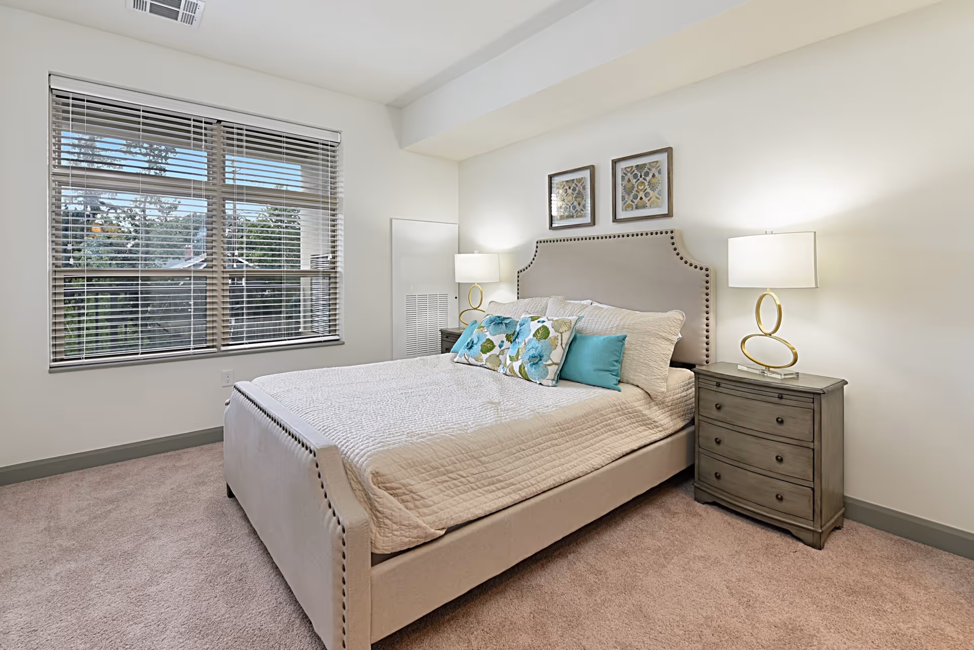 Bright bedroom featuring an upholstered bed with decorative pillows, two bedside tables with lamps, framed art, and a large window.
