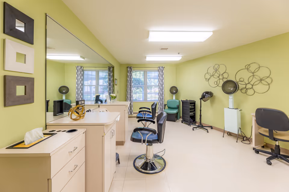 A bright salon room with light green walls, featuring two black salon chairs in front of a large mirror, hair drying stations with hooded dryers, a small desk with a rolling chair, and a window with patterned curtains letting in natural light.