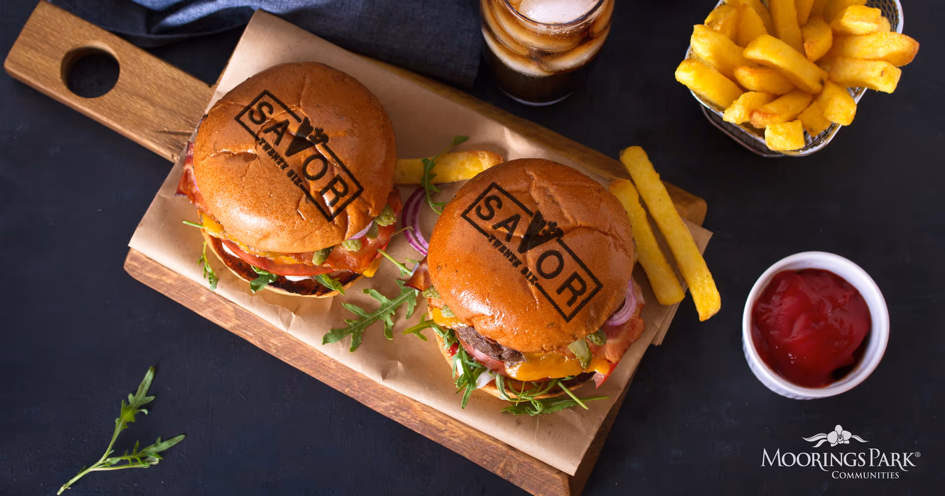 Two cheeseburgers with lettuce, tomato, and onions on wooden serving board with fries, a small bowl of ketchup, and a glass of iced drink on a dark surface.