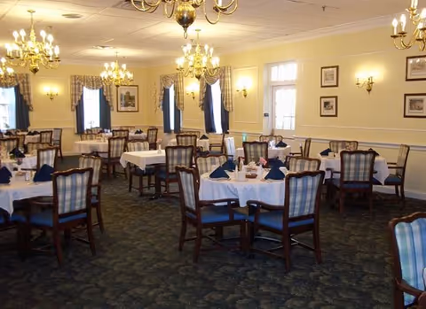 A spacious dining room with multiple round tables covered with white tablecloths and set with blue napkins. The room features wooden chairs with blue and white plaid upholstery, chandeliers hanging from the ceiling, framed pictures on the walls, and windows with blue curtains and valances.