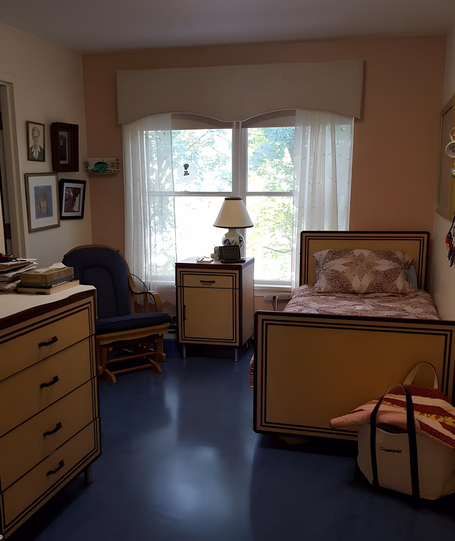 Sunlit single-occupancy bedroom with a bed, nightstand and lamp, dresser and armchair in front of a large window.