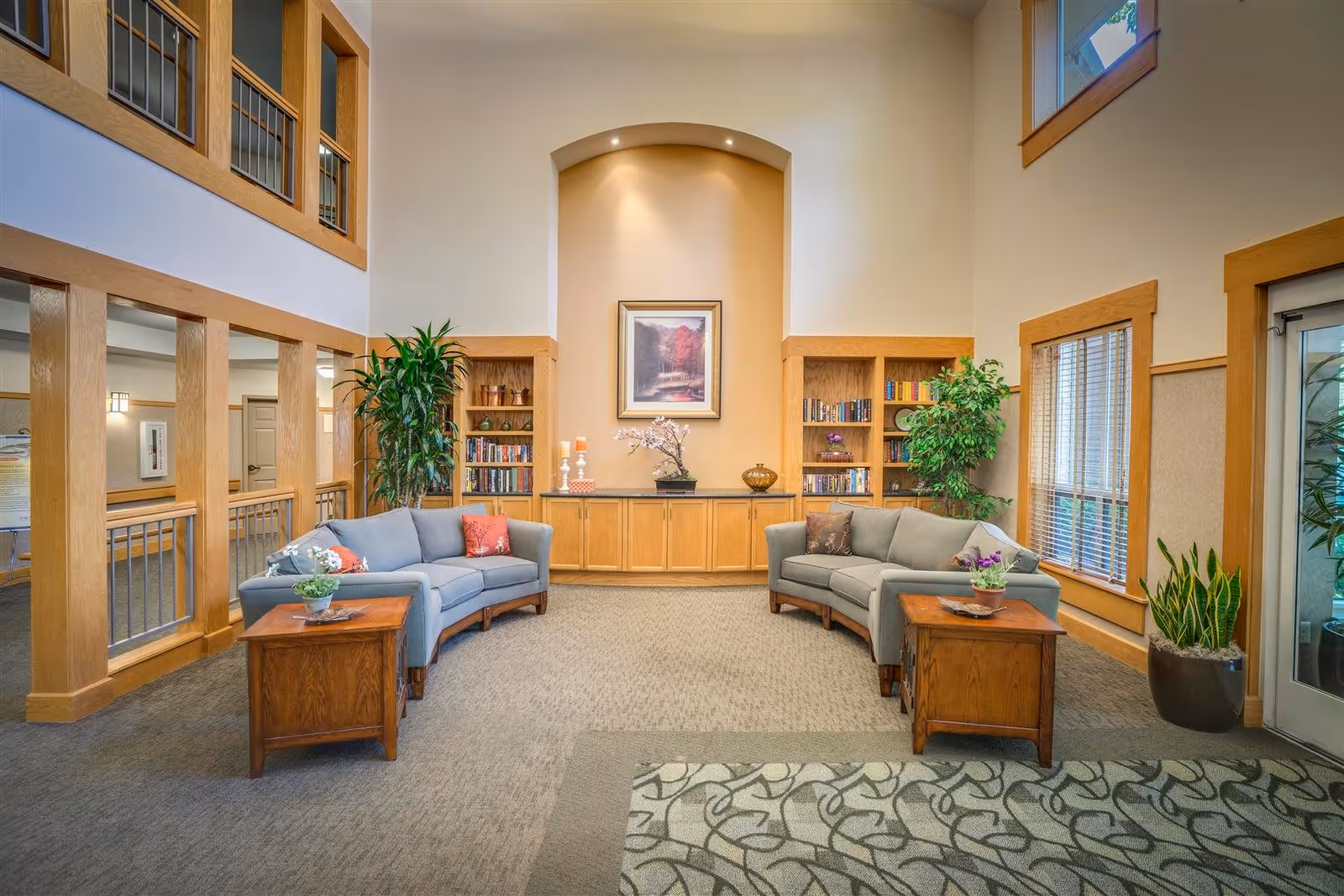 A bright and spacious common area with two curved gray sofas facing each other, each with decorative pillows. Between the sofas are wooden side tables with potted plants. The back wall features built-in wooden bookshelves filled with books and decorative items, and a framed picture of a scenic landscape hangs in a recessed, well-lit alcove. Large windows with wooden trim allow natural light to enter the room, and there are several green plants placed around the space. The carpet has a neutral tone with a patterned area rug in the foreground.