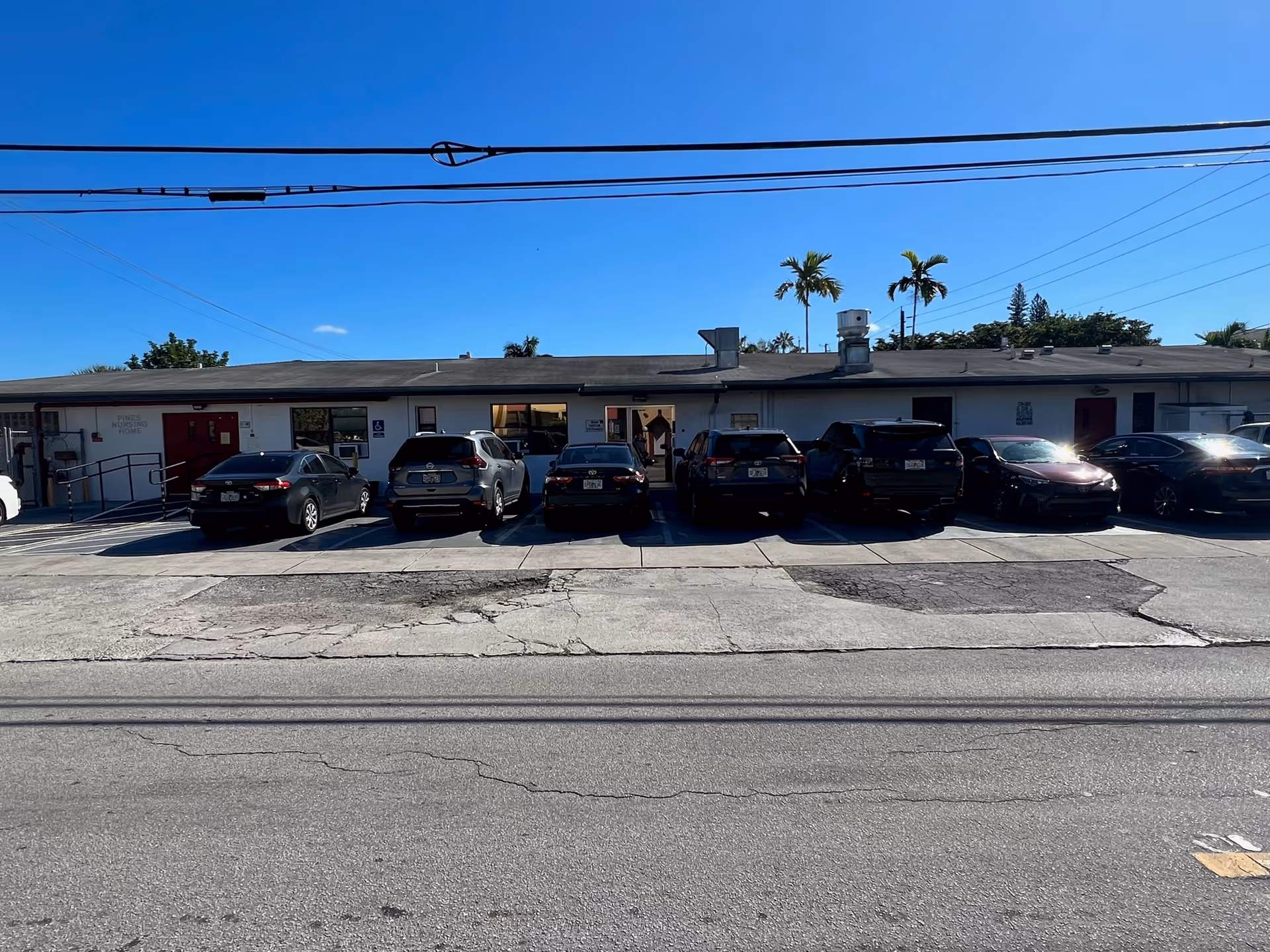 Single-story nursing home building with a row of parked cars in front, palm trees behind, and a clear blue sky.