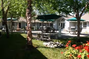 Sunny courtyard with trees, lawn, patio tables and umbrellas in front of a single-story retirement community building.