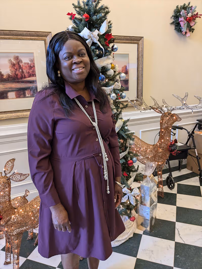A woman wearing a purple dress and a long pearl necklace stands smiling in front of a decorated Christmas tree with ornaments and ribbons. Next to her are two lighted reindeer decorations and wrapped presents. The background includes framed artwork on the wall and a walker.