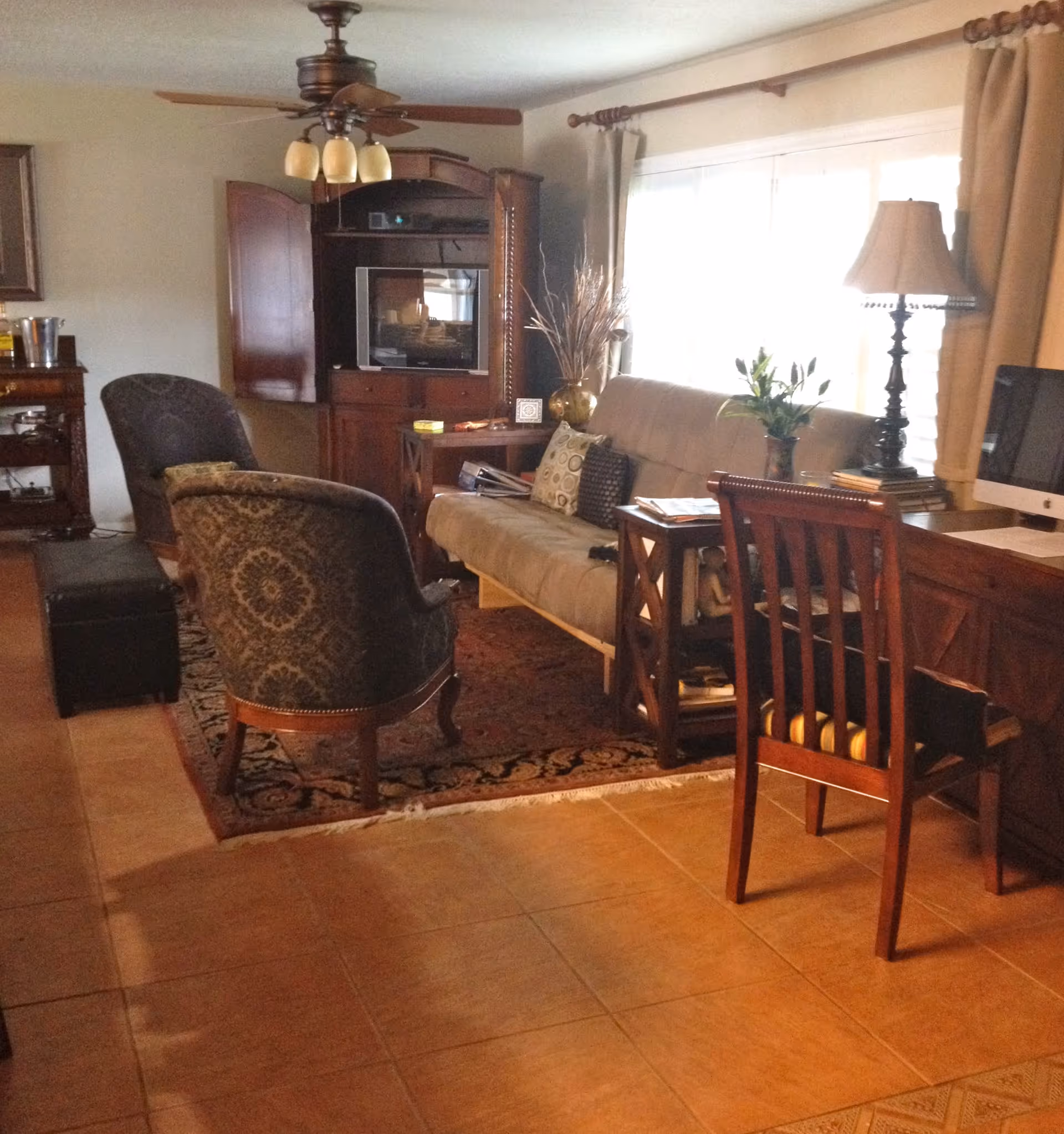A cozy living room with a beige sofa adorned with patterned pillows, two upholstered armchairs, a wooden entertainment center with a TV, a ceiling fan with lights, a wooden desk with a chair and a computer, a floor lamp, and large windows with curtains allowing natural light to fill the room.