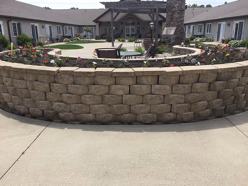 Courtyard with a curved raised stone planter filled with flowers, patio seating, and a pergola surrounded by single-story residential buildings.