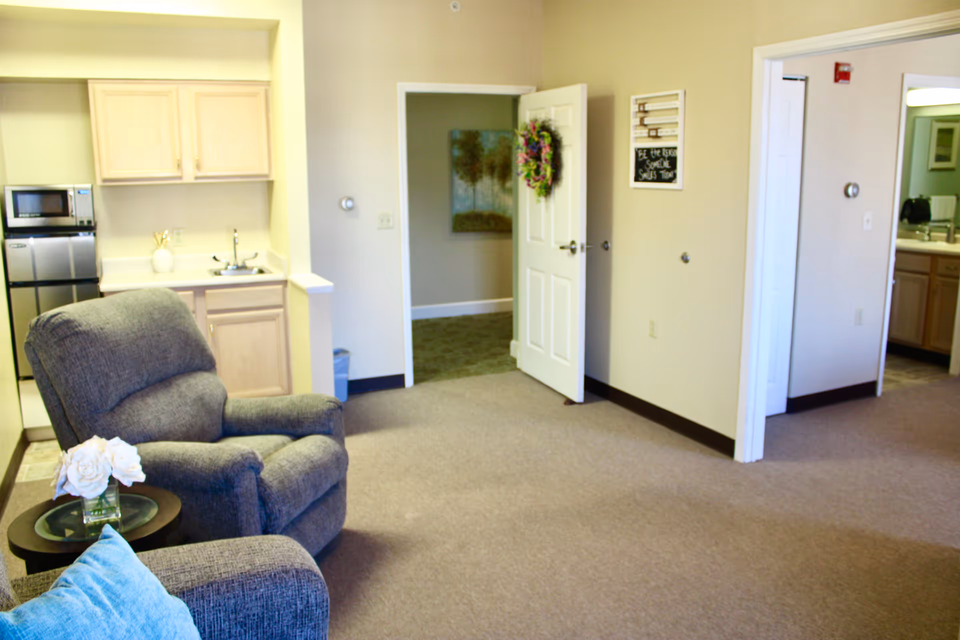 A cozy living room area with a gray upholstered recliner chair and a small round side table holding a vase with white flowers. In the background, there is a small kitchenette with light wood cabinets, a sink, a microwave, and a mini refrigerator. Two open doorways lead to other rooms, one decorated with a wreath on the door and a painting on the wall.