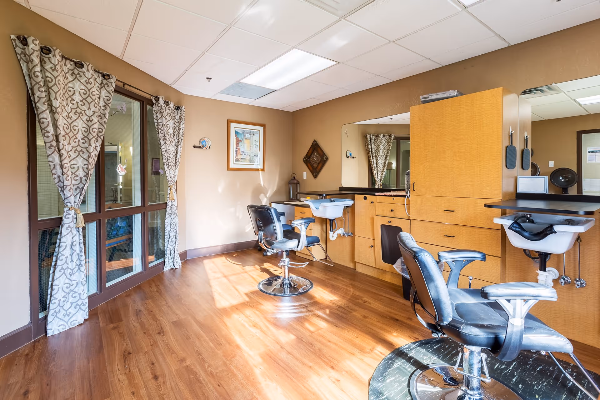 Interior of a salon room with two black salon chairs, wooden cabinetry with sinks, large mirrors, and patterned curtains on windows letting in natural light.