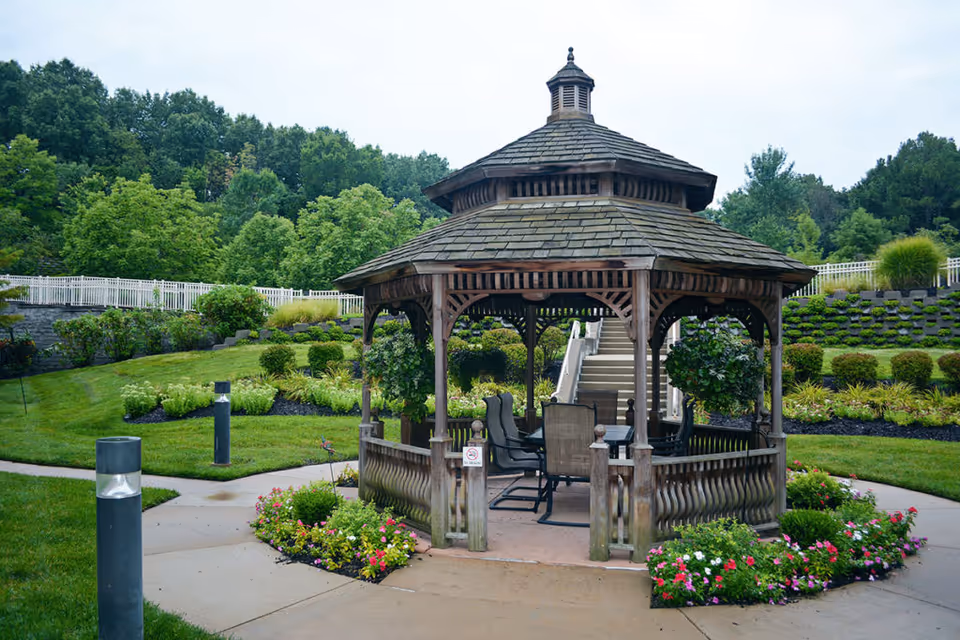 A wooden gazebo with a shingled roof situated in a landscaped garden area with green grass, colorful flowers, and shrubs. The gazebo contains several chairs and a table inside. There are stairs and a white fence in the background, with trees and bushes surrounding the area.