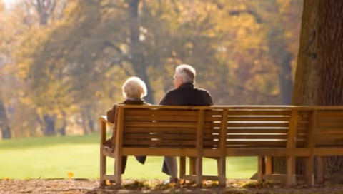 Two elderly people sit on a wooden bench in a park surrounded by autumn trees.