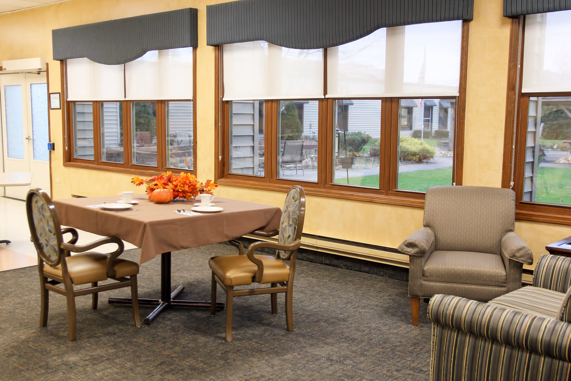 A cozy dining area in a senior living facility with a table covered by a brown tablecloth, set with white plates, cups, and silverware. The table has a centerpiece of autumn leaves and a small pumpkin. There are two cushioned chairs with armrests around the table. Large windows with white blinds and blue valances let in natural light, and comfortable upholstered armchairs are visible nearby. Outside, a garden and patio area can be seen through the windows.