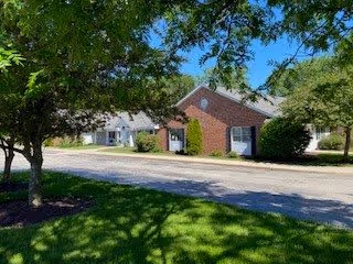 A sunny outdoor scene showing a paved road with green grass and trees casting shadows. On the right side, there is a single-story brick building with white trim and windows, surrounded by bushes and trees under a clear blue sky.