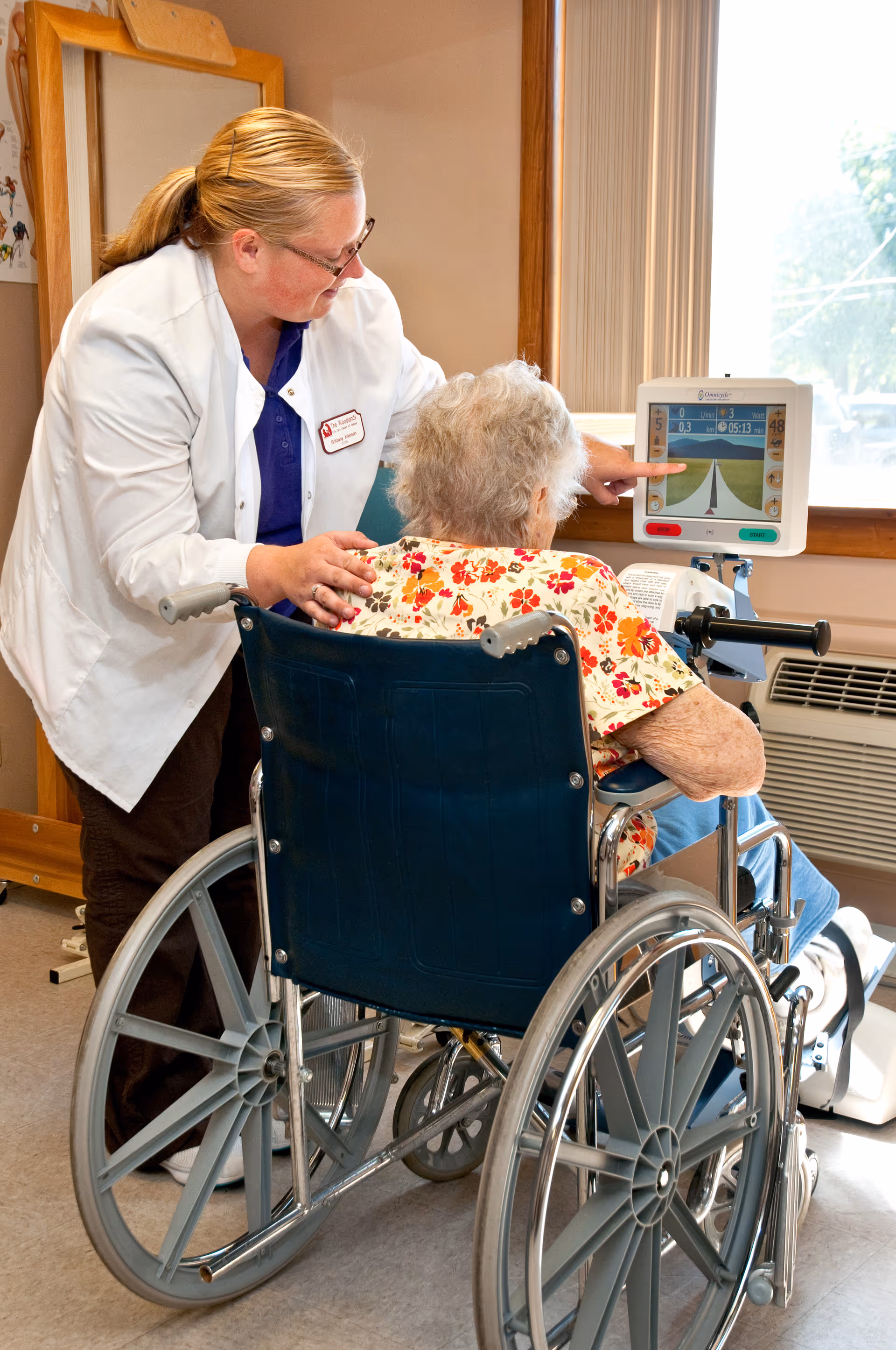 A healthcare worker in a white coat assists an elderly woman in a wheelchair as they look at a screen displaying a virtual road and exercise data in a well-lit room with a window.