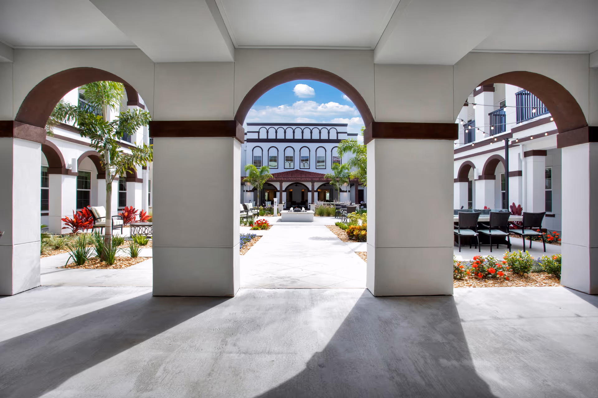 View through three large archways of a courtyard area at The Blake at LPGA, featuring outdoor seating, landscaped plants, palm trees, and a white building with dark trim under a blue sky with clouds.
