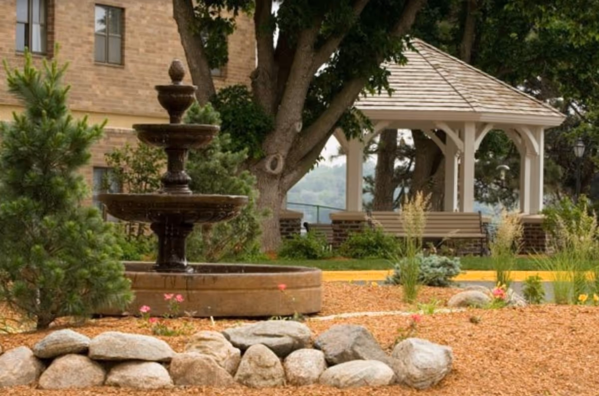 Outdoor garden area at Keystone Villas Senior Living featuring a tiered water fountain surrounded by rocks and small plants, with a white gazebo and benches in the background under large trees.