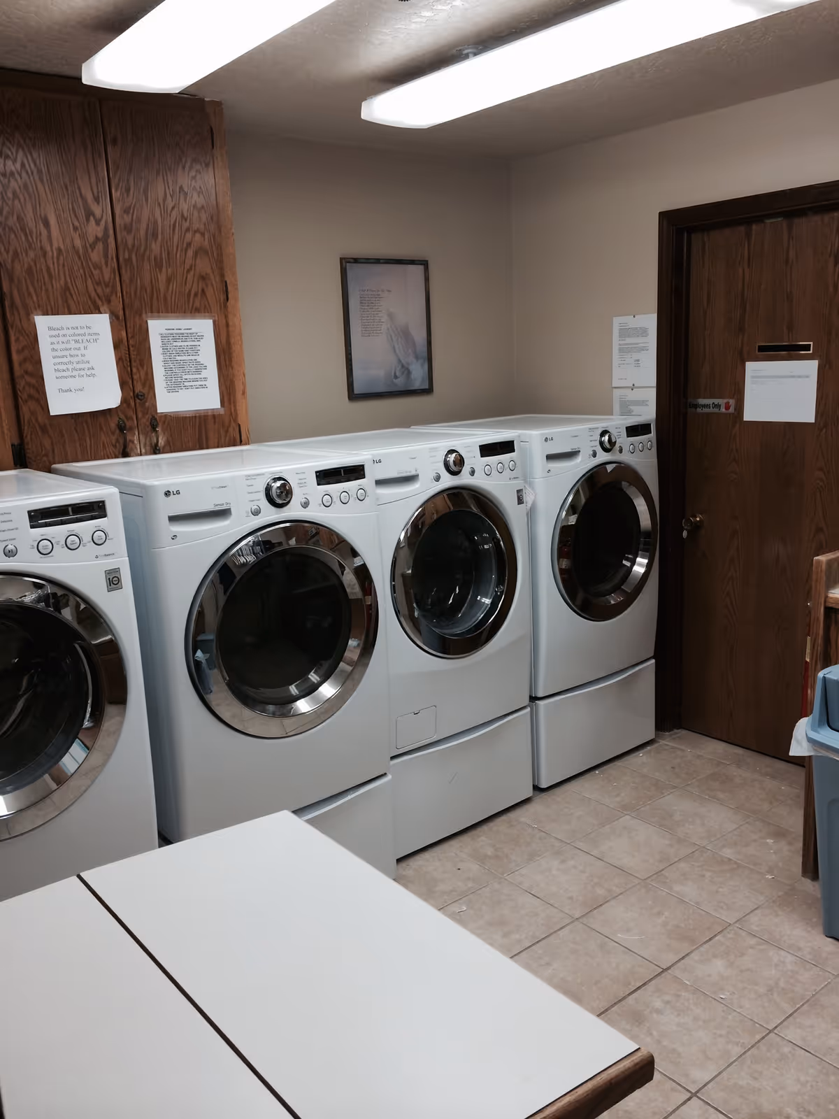 Laundry room showing multiple front-loading washers and dryers, a folding table, wooden cabinets, and tiled floor.
