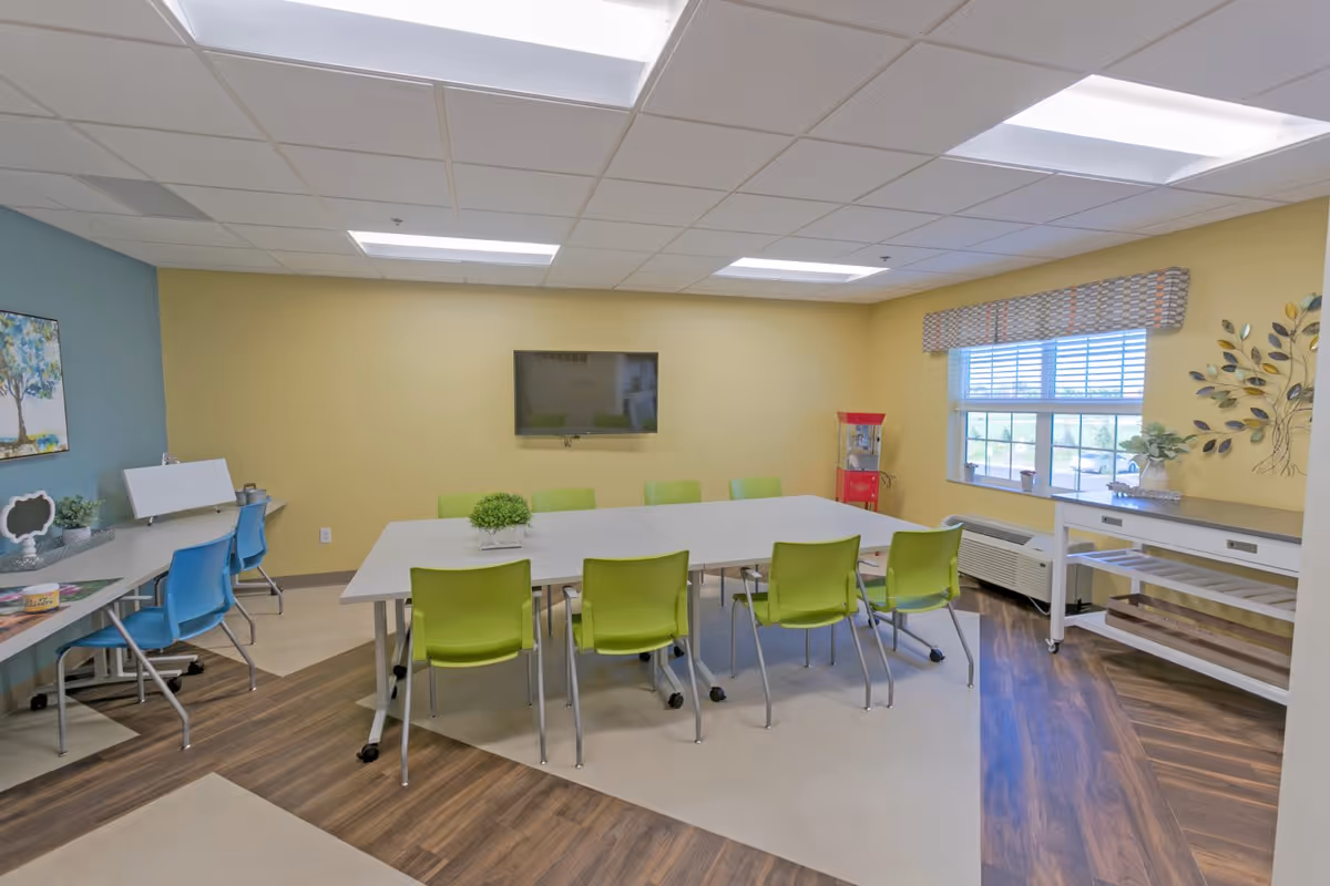 A brightly lit activity or meeting room with a large white table surrounded by green chairs. There are blue chairs along a side desk with some decorative items and a painting on the wall. A flat-screen TV is mounted on a yellow wall, and a popcorn machine is placed near a window with blinds and a patterned valance. The floor has a mix of wood and light-colored tiles, and there is a white console table with plants and decor near the window.