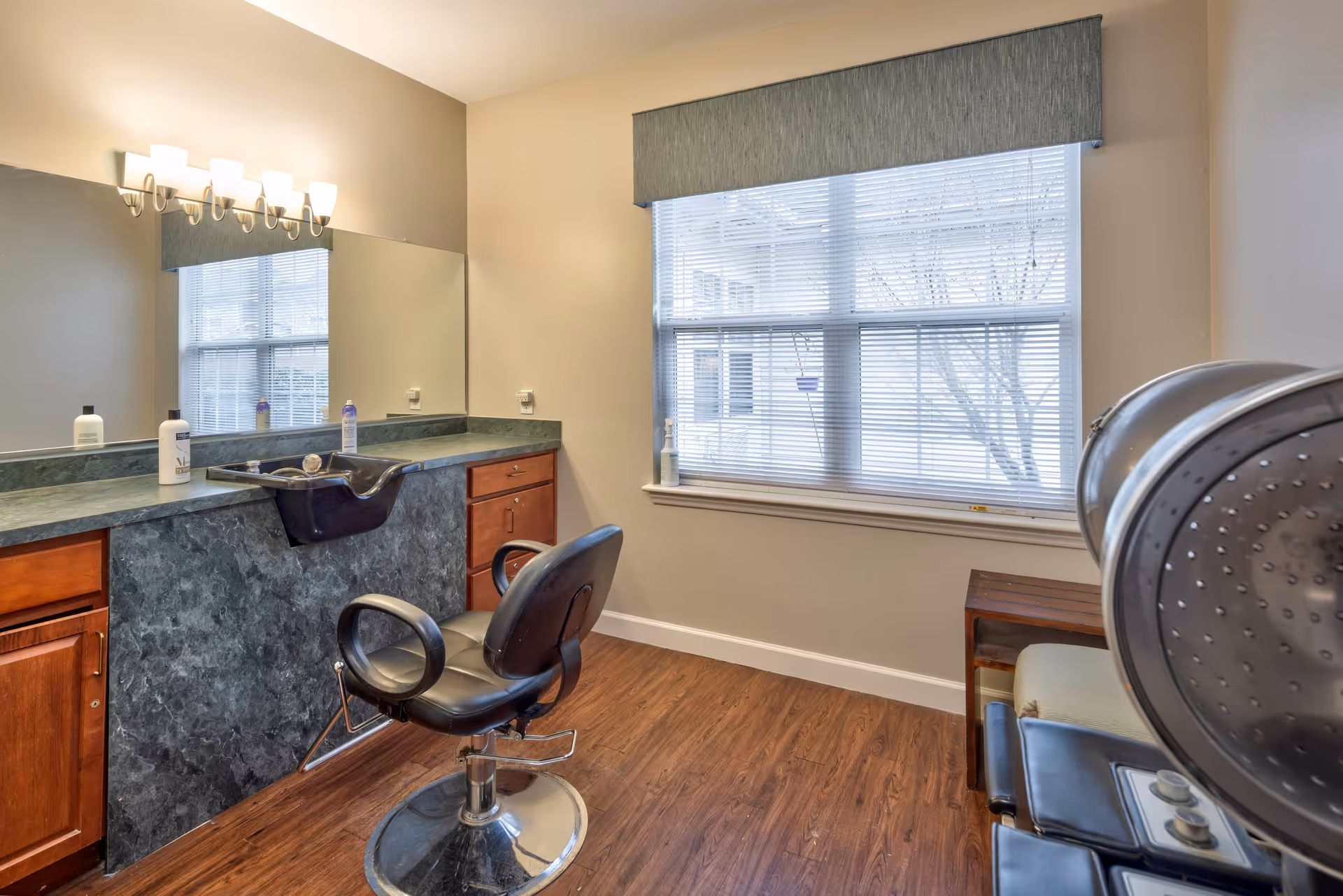 Interior view of a hair salon area in a senior living facility with a black salon chair in front of a counter with a built-in sink, bottles of hair products on the counter, a large mirror on the wall, a window with blinds and a valance, and a hair dryer chair on the right side.