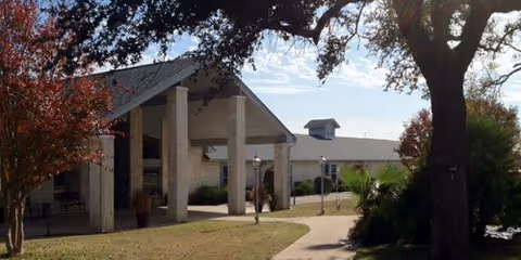 Covered entrance of a senior living building with tall columns, a walkway, and surrounding trees and shrubs.