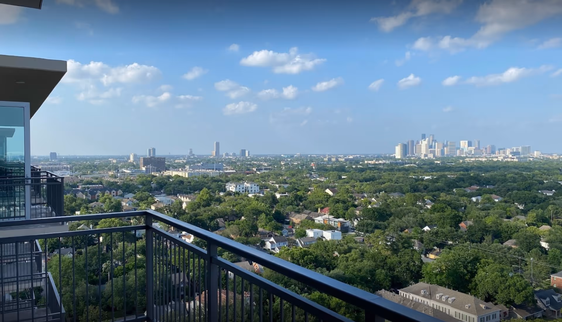View from a balcony overlooking a cityscape with many trees and buildings under a blue sky with scattered clouds.
